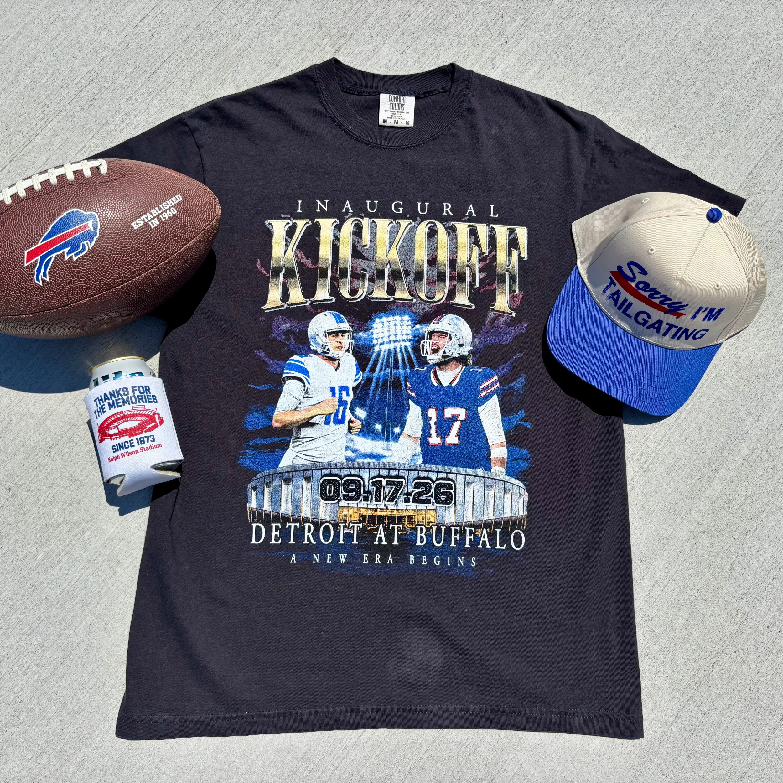 A navy blue graphic t-shirt featuring an inaugural kickoff design for Detroit at Buffalo, styled in a flat lay with a brown football, a can cooler, and a blue and white cap on a gray concrete background.