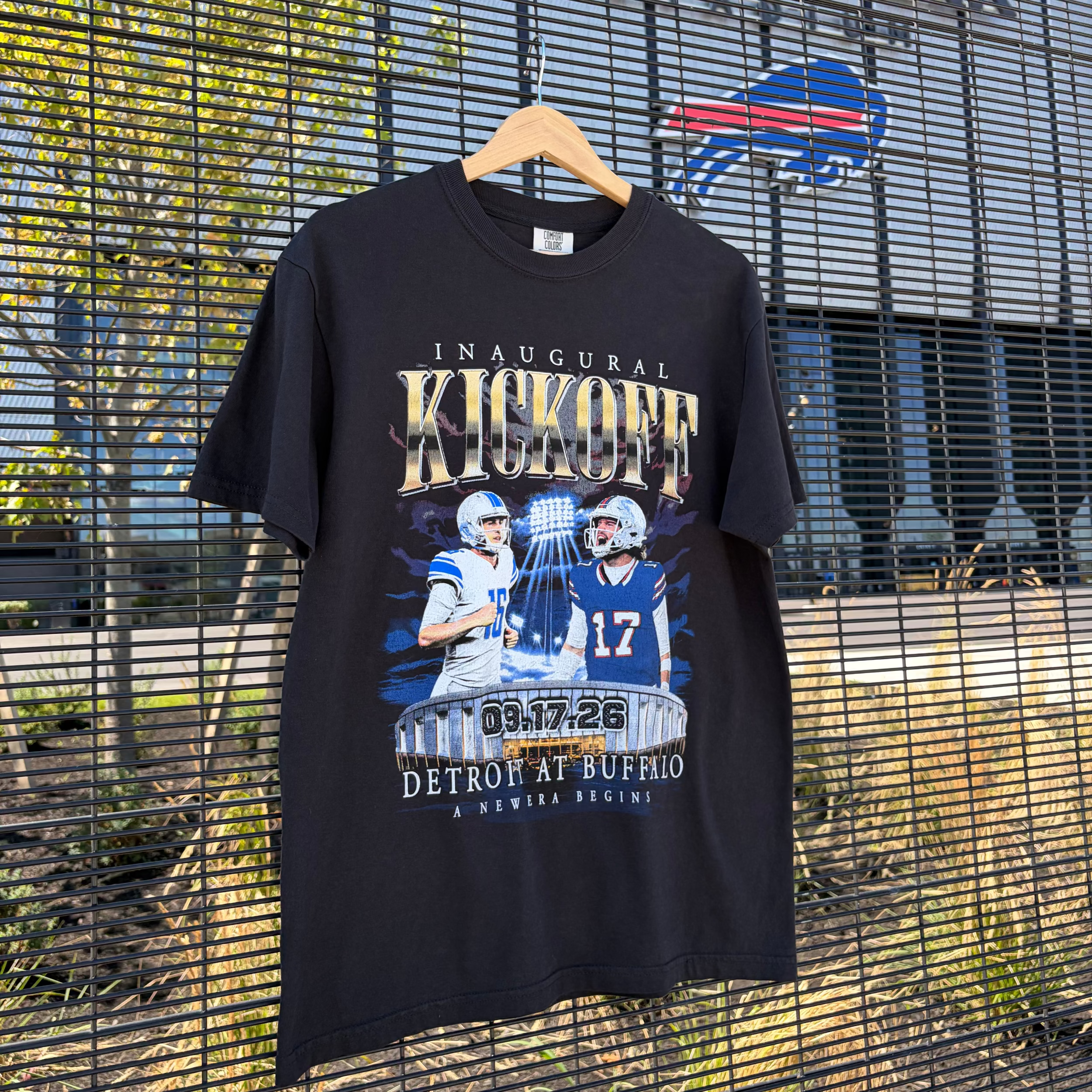 A black short-sleeve graphic T-shirt hanging on a wooden hanger in front of a metal fence and building facade. The shirt features a large football-themed graphic with the words 'Inaugural Kickoff', player photos, and 'Detroit at Buffalo' text.